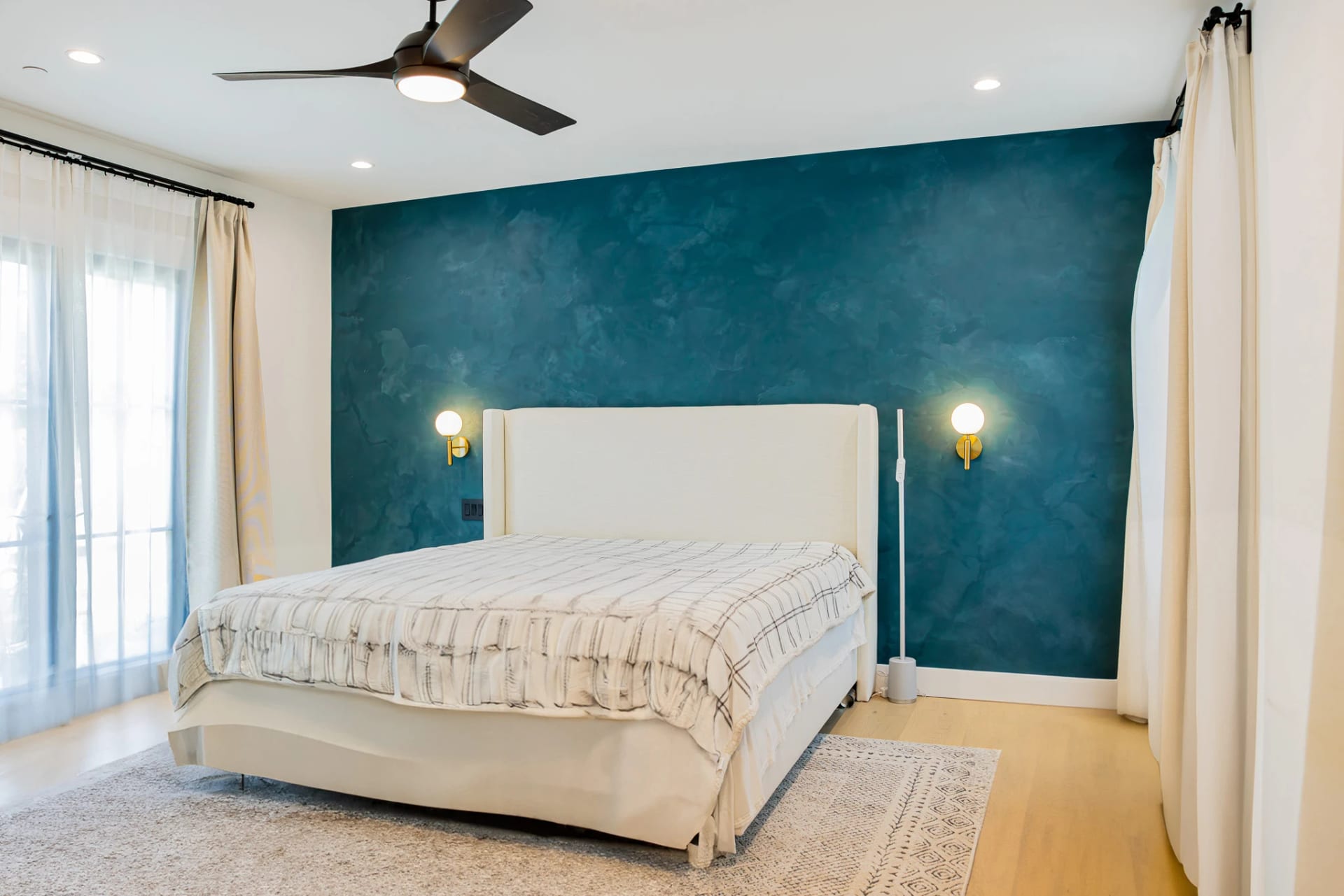 Bedroom with a deep blue plaster accent wall, wall sconces and a ceiling fan