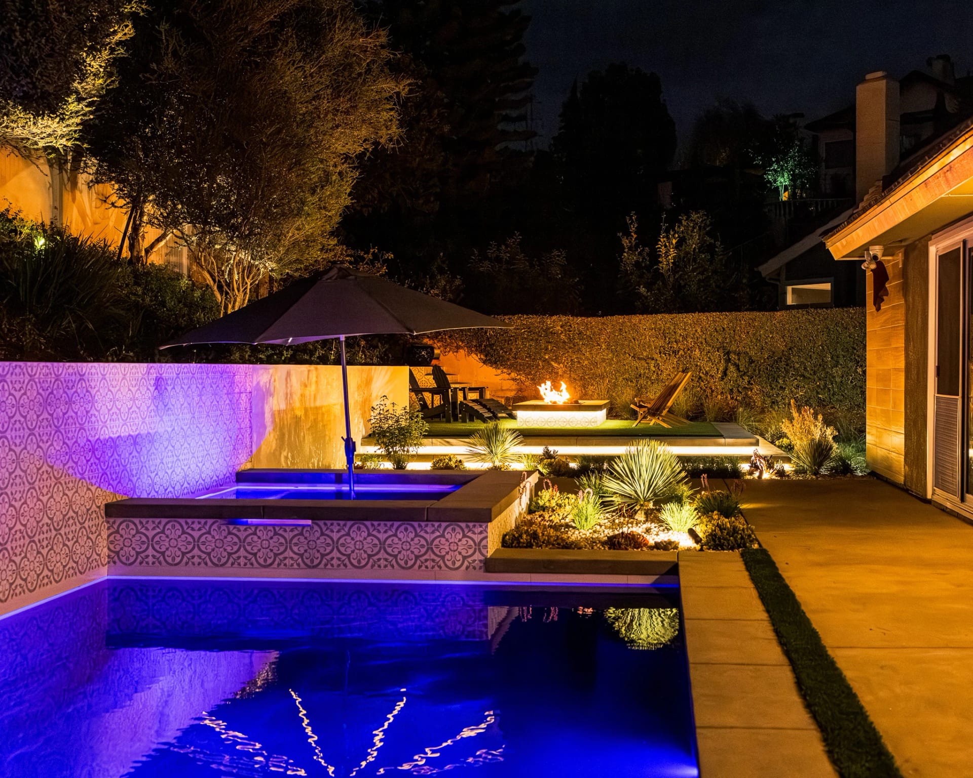 Spa and pool glowing blue at night with a fire feature behind